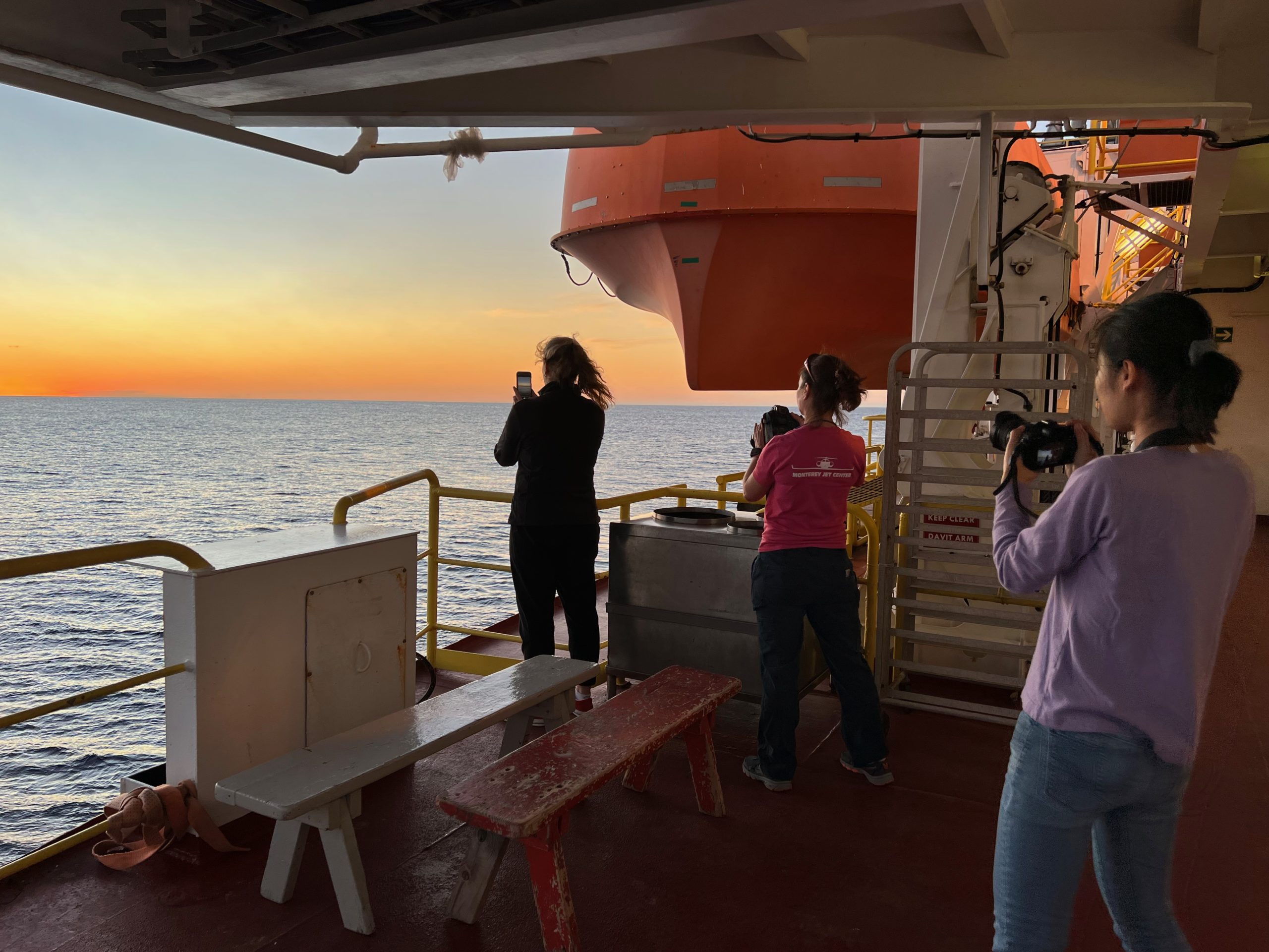 Three female scientists at sea taking a photo of the sunset