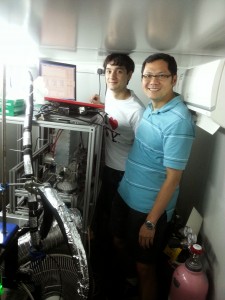 Berto Lee (left) and Chak Chan (right) stand in a shelter in Hong Kong’s Mong Kok next to the Aerodyne High Resolution Aerosol Mass Spectrometer, used to sample airborne particulate matter.  Credit: Chak Chan