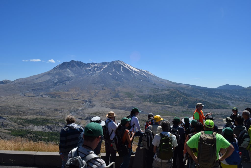 On a clear, sunlight afternoon, a crowd listens to a speaker on the platform overlooking the devastated zone north of Mount St. Helens volcano. The deep crater from the 1980 eruption is clearly visible in the distance.