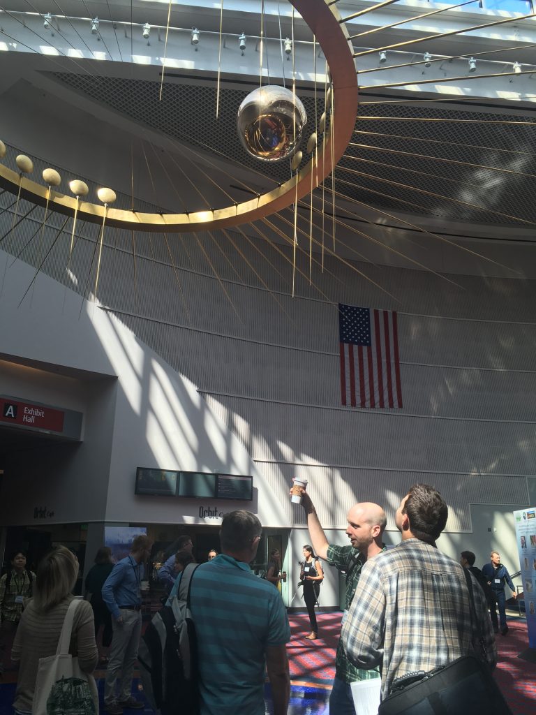 Scientists stand below a suspended Foucault Pendulum art piece in a sunlit lobby of the Oregon Convention Center.